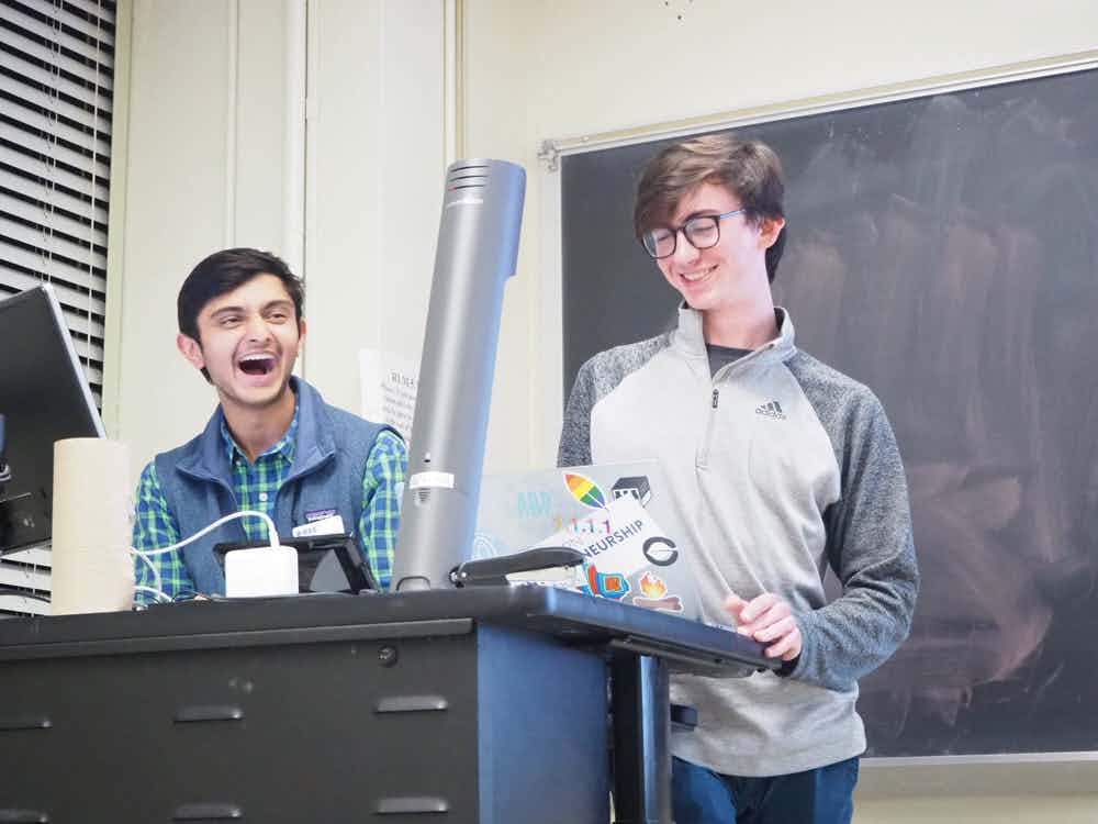 2 IEEE members in a classroom podium laugh while looking at a laptop